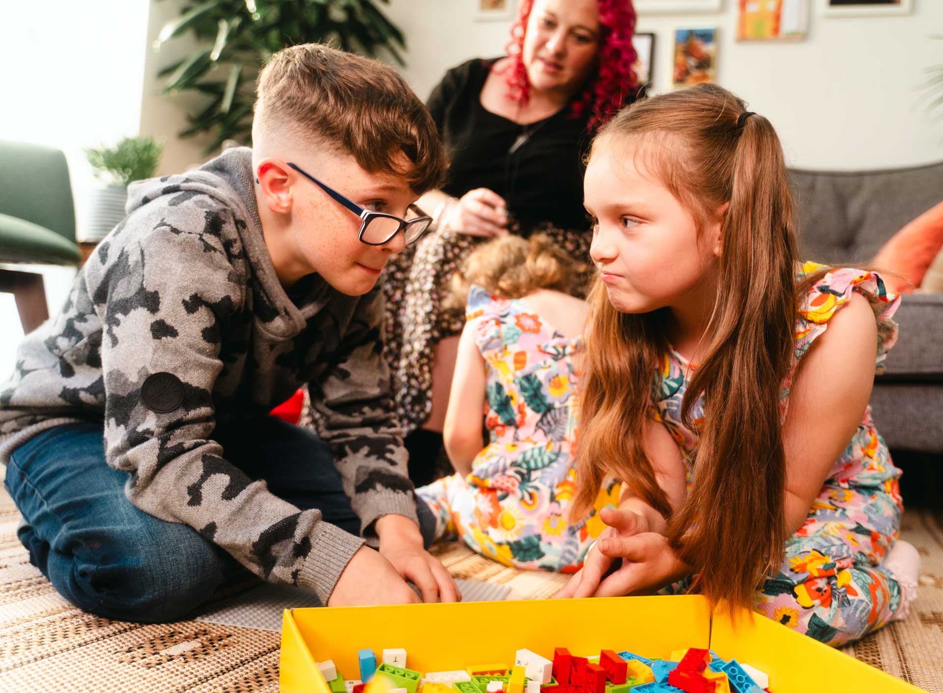 Young boy with vision impairment and his sister looking at each other beside an open box of LEGO® Braille Bricks. Mum watches in the background. The young boy has a pale complexion and short brown hair, who wears black framed glasses. His mum has a pale complexion and vibrant pink curly hair down to her shoulders. His sister has a pale complexion and light brown hair.